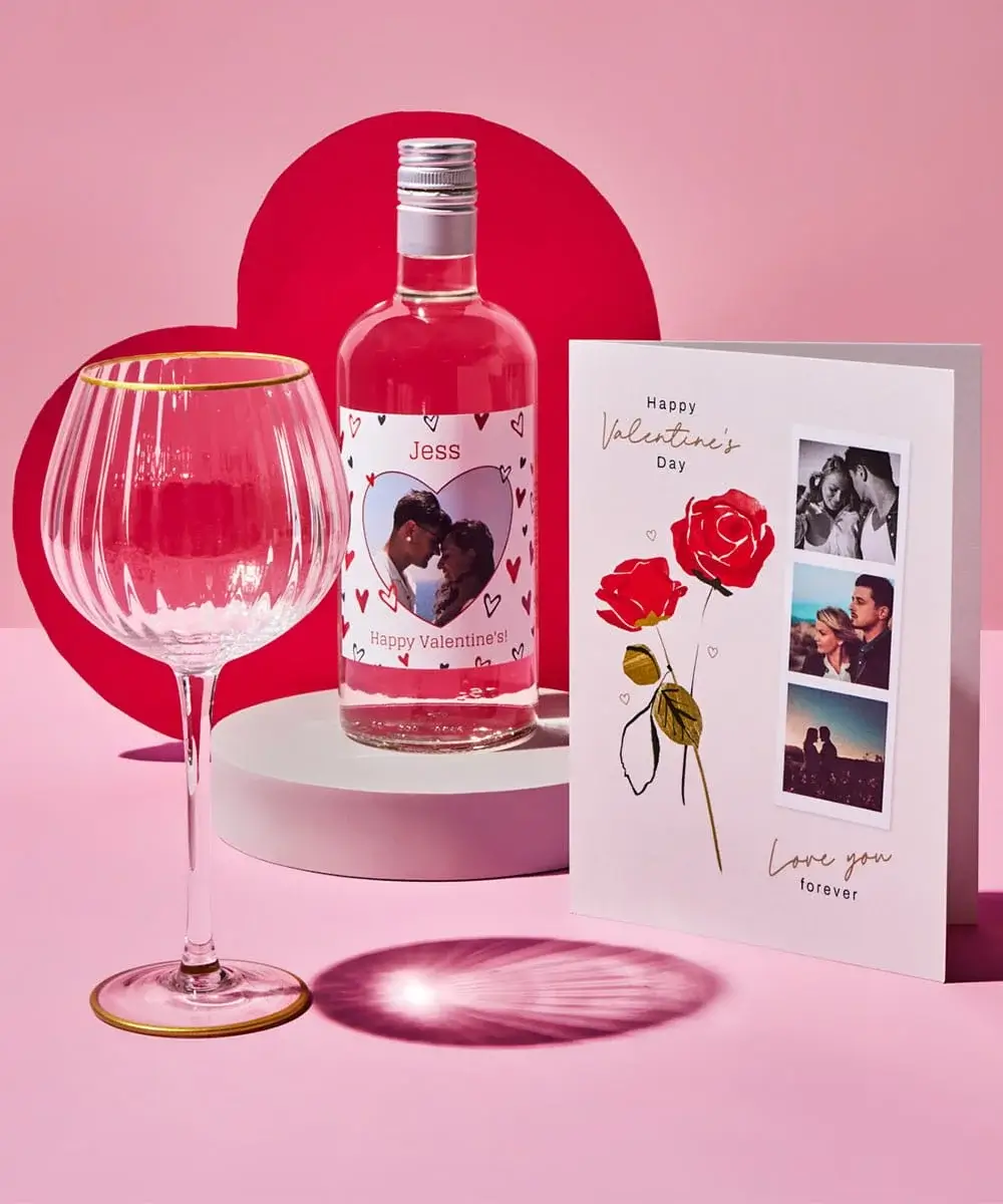 Valentine’s Day gift set displayed on a pink background with red heart-shaped props. It includes a clear wine glass with a gold rim, a bottle of rosé wine featuring a custom label with heart illustrations and the text Jess Happy Valentine’s, and a greeting card decorated with two red roses and three photo slots. The card reads Happy Valentine’s Day and Love you forever.